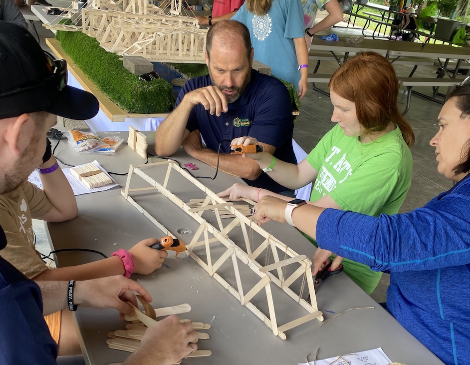 People sit around a table looking at a small bridge built with popsicle sticks.