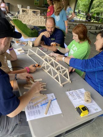 People sit around a table looking at a small bridge built with popsicle sticks.